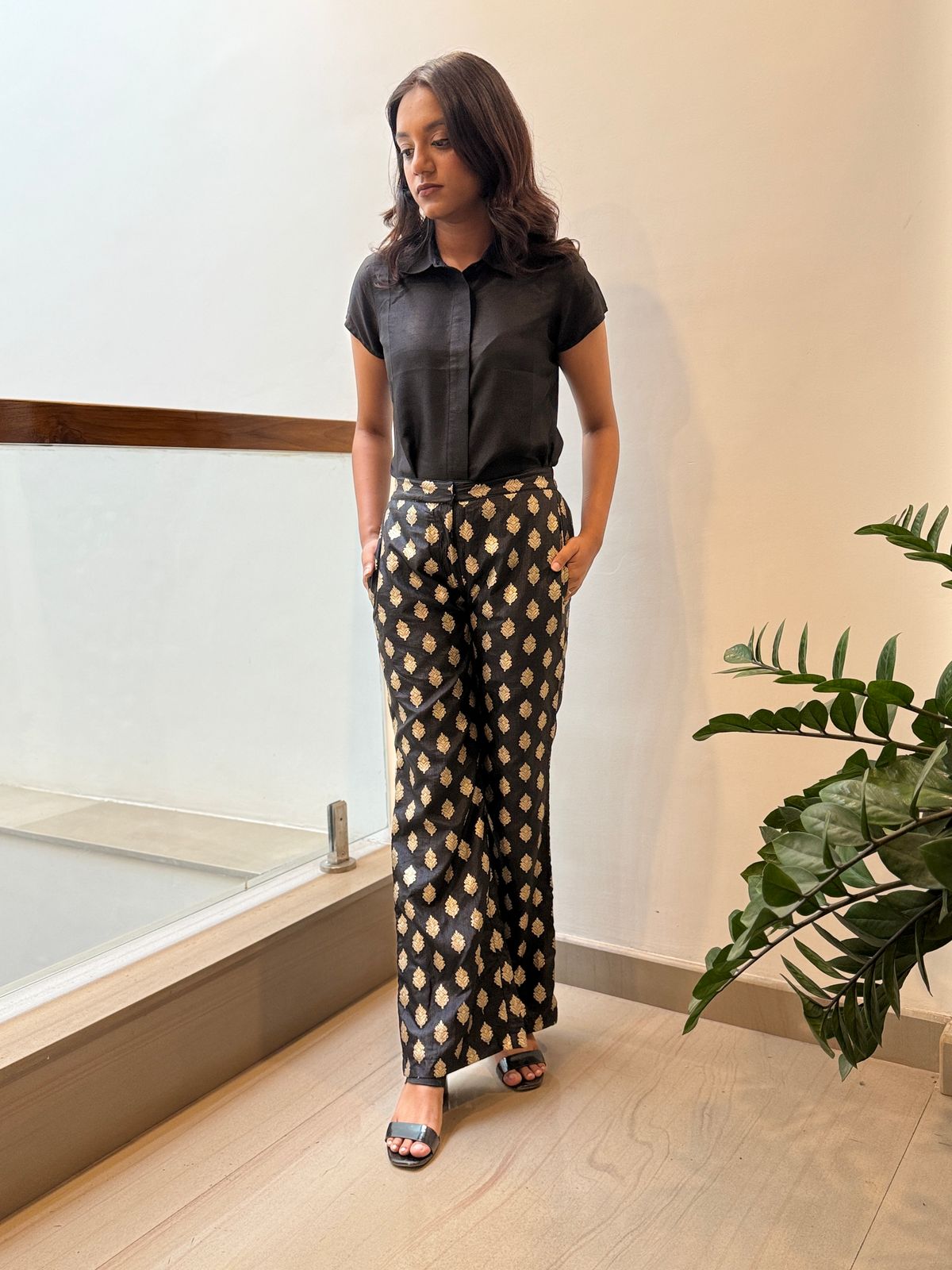Woman in black shirt and patterned skirt standing indoors next to a window with a plant.
